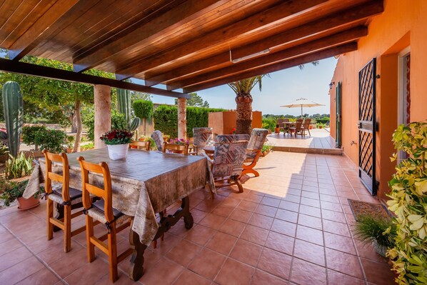 An outdoor patio with a wooden table and chairs, surrounded by plants and a view of the landscape. - With pool in a quiet location - Villa Ses Salines (Ses Salines)