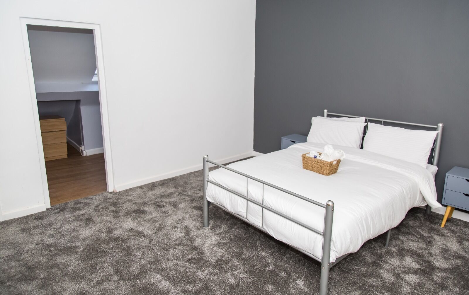 Een slaapkamer met een metalen bedframe, witte beddengoed, een rieten mandje op het bed en een grijs vloerkleed.
