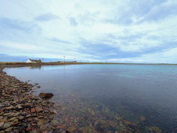 A calm coastal scene with a pebble beach, a white building, and distant power lines. - Mingro is a unique beachside cottage that serves as a retreat from modern life. (Stronsay, near Whitehall)
