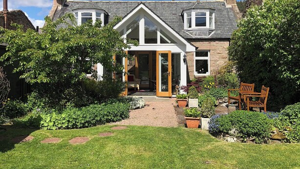 Une maison à deux étages avec un porche à l’avant, une véranda et un jardin agrémenté d’un banc.