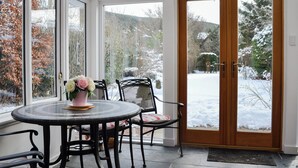 Un coin repas avec une table ronde et des chaises, un vase de fleurs, et une vue sur un paysage enneigé grâce à de grandes fenêtres et une porte en bois.