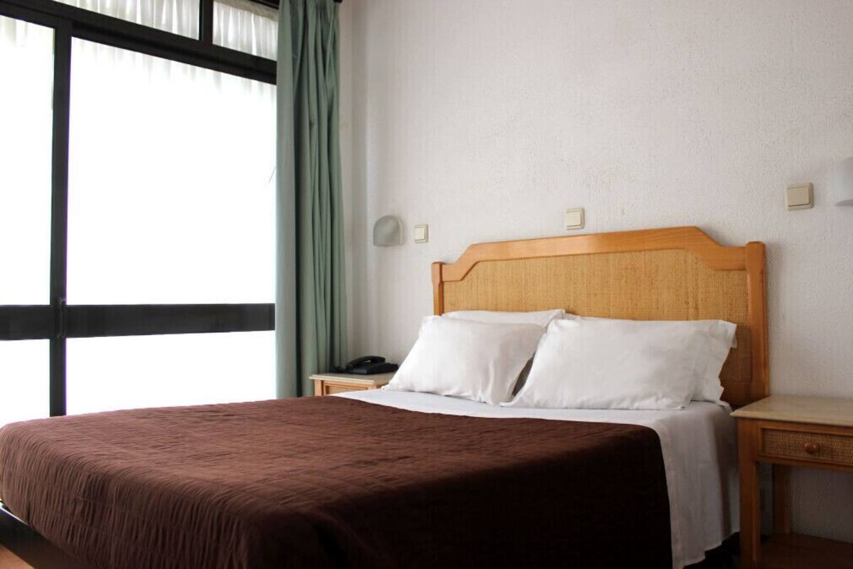 A hotel room with a large bed, a wooden headboard, a bedside table with a lamp, and a window with curtains.