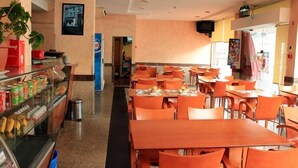 Uma cafeteria com mesas e cadeiras laranja, um balcão de vidro com lanches e uma geladeira da Pepsi.