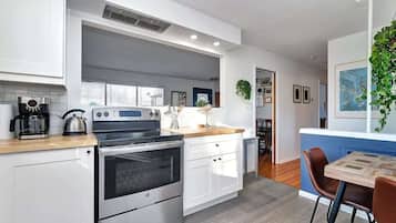A modern kitchen with white cabinets, a built-in oven, and a coffee maker. There is a dining area with a wooden table and chairs.