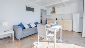 Una sala de estar moderna con un sofá gris, una mesa de comedor y una zona de cocina con armarios y electrodomésticos blancos.