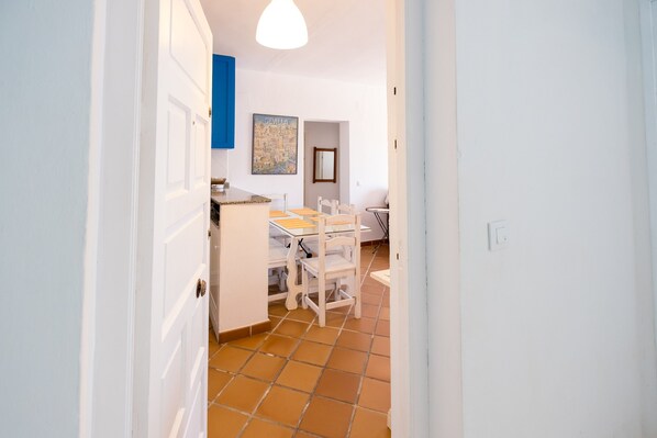 Un pasillo que conduce a una cocina con armarios blancos, suelo de baldosas y un comedor con sillas y mesa blancos.