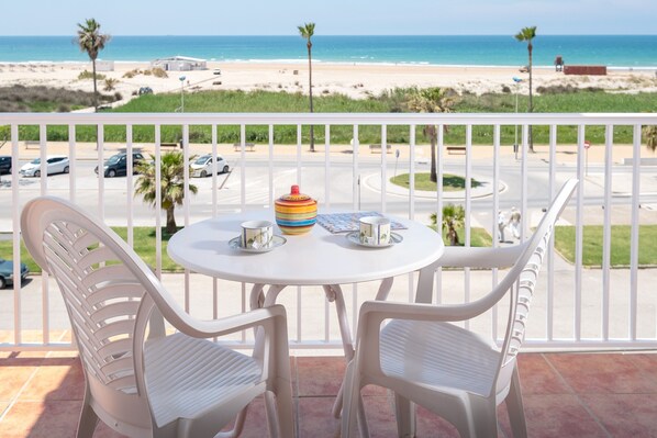 Un balcón con sillas y mesa blancos, con vistas a una playa con palmeras y un cielo despejado.