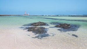 A beach with clear water, rocks, and a distant lighthouse.