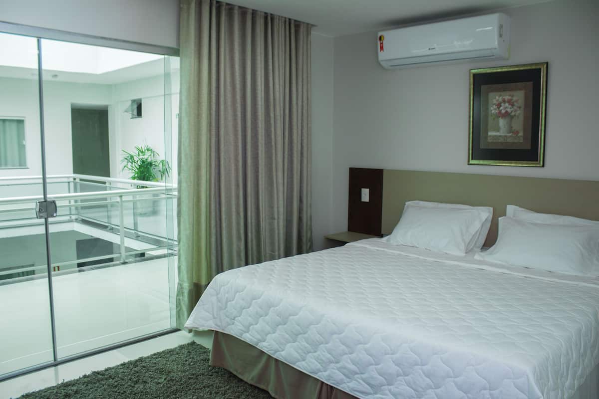 A hotel room with a bed, a nightstand, a framed picture, a window with curtains, and a balcony with a railing.