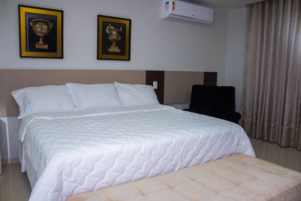 A hotel room with a large bed, two framed trophies on the wall, an air conditioner, and a chair.