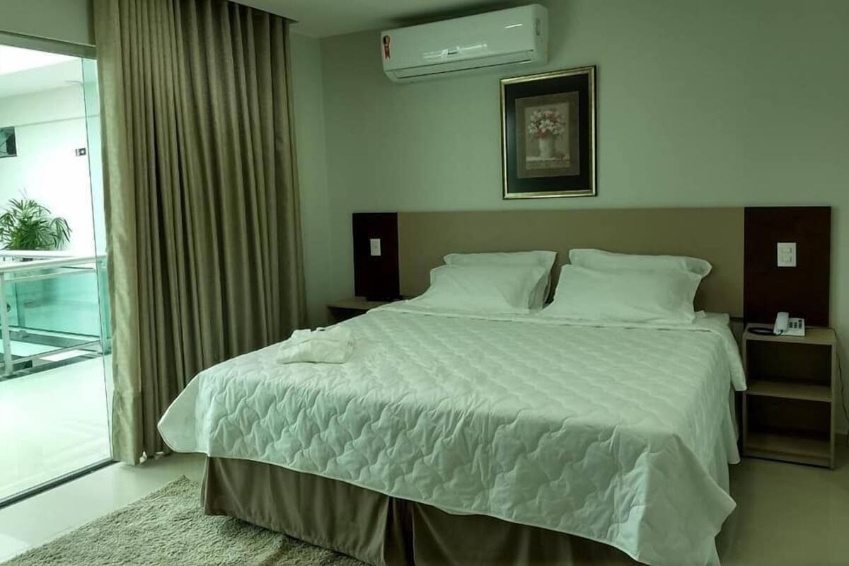 A hotel room with a bed, a bedside table, a nightstand, an air conditioning unit, a framed picture on the wall, and a view of the outside balcony.