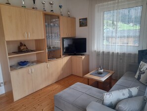 Ein Wohnzimmer mit einer Couch, einem Fernseher, einer Vitrine mit Pokalen und einem Fenster mit Blick.