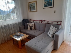 Un salon avec un canapé gris, une petite table en bois et une fenêtre ornée de rideaux légers.