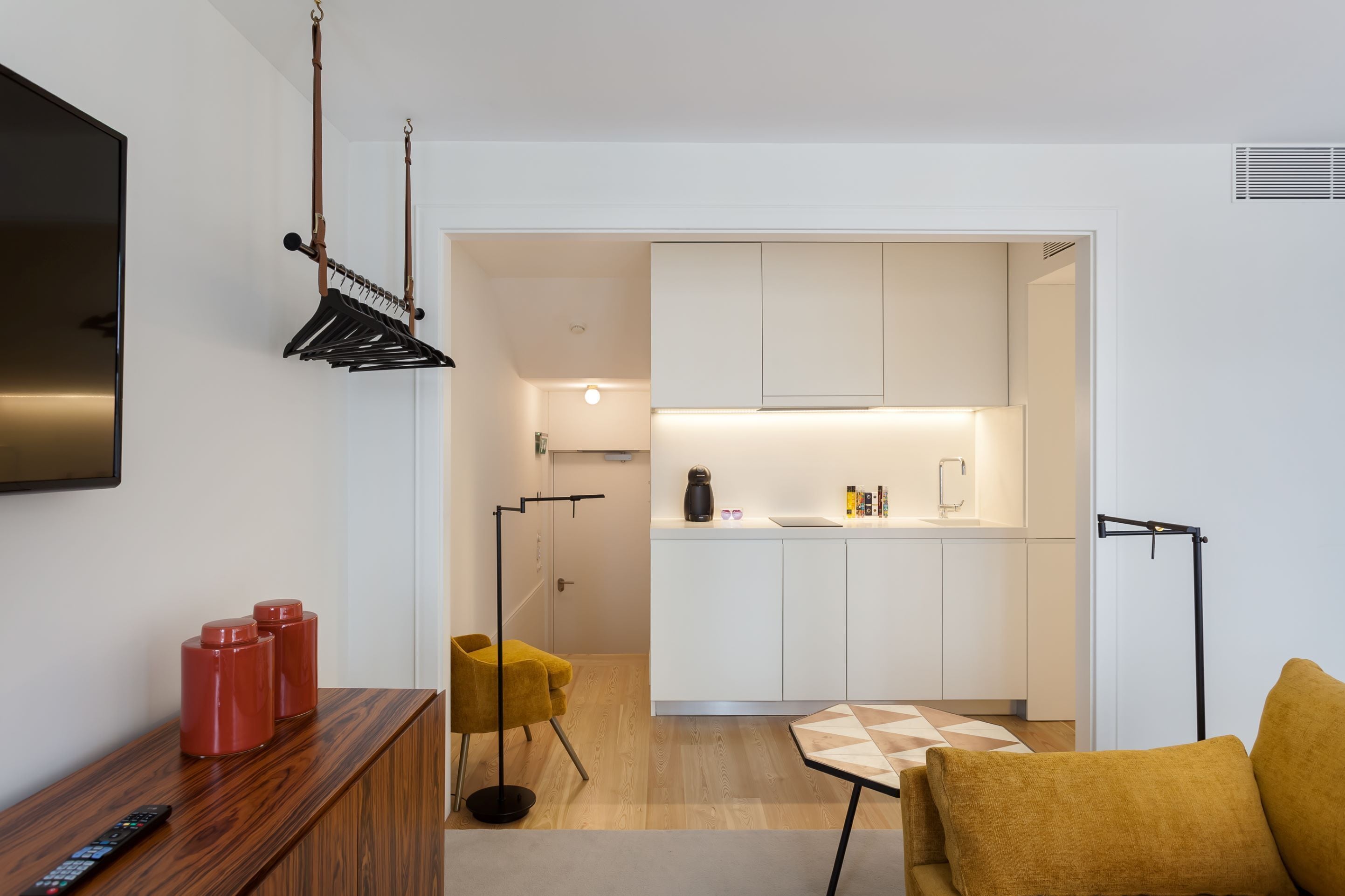 Una sala de estar moderna con un mueble bajo para la televisión de madera, un sillón amarillo y un reposapiés con estampado geométrico. Se ve una cocina con armarios blancos y encimera negra a través de un umbral abierto.