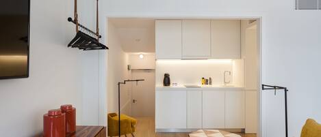 Una sala de estar moderna con un mueble bajo para la televisión de madera, un sillón amarillo y un reposapiés con estampado geométrico. Se ve una cocina con armarios blancos y encimera negra a través de un umbral abierto.