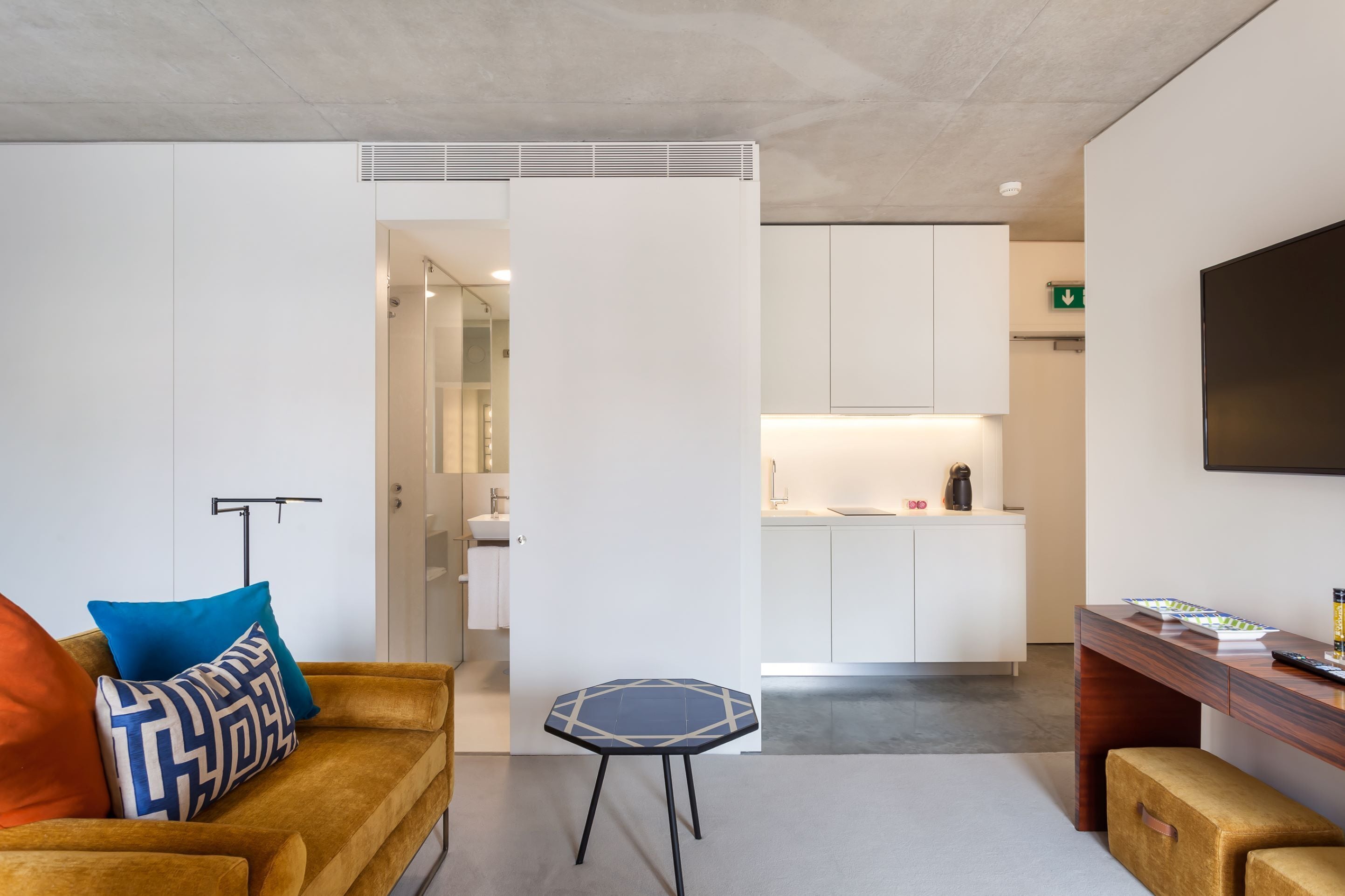 A modern hotel room with a sofa, a small table, a TV, and a kitchenette in the background.