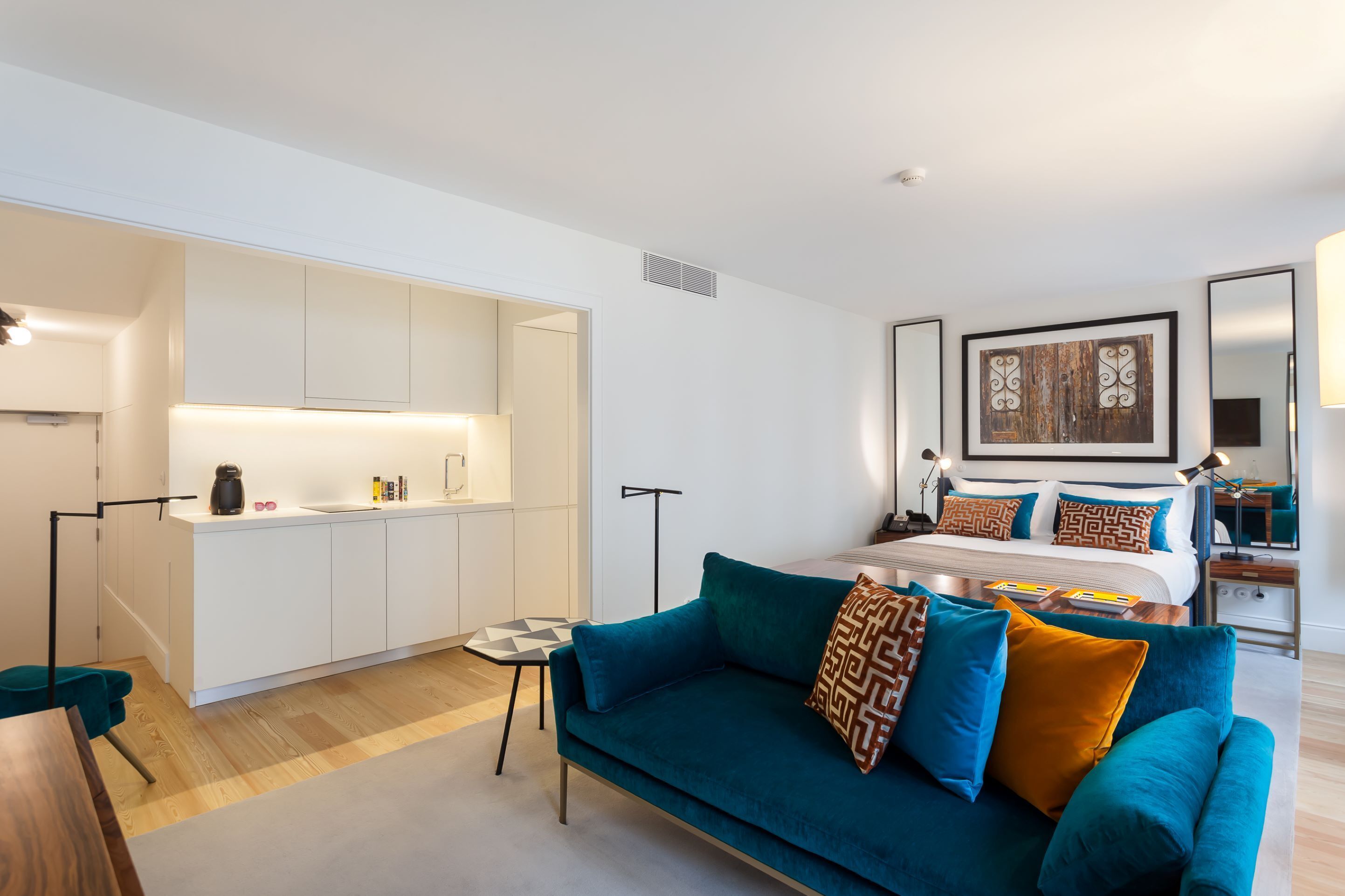 A modern hotel room with a kitchenette, a large bed with patterned pillows, a teal sofa, and a small side table.