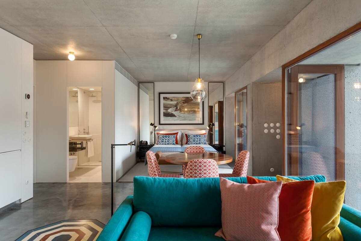 A modern hotel room with a teal sofa, a bed with patterned pillows, a dining area with a round table, and a large window with a view.