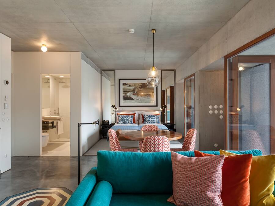 A modern hotel room with a teal sofa, a bed with patterned pillows, a dining area with a round table, and a large window with a view.