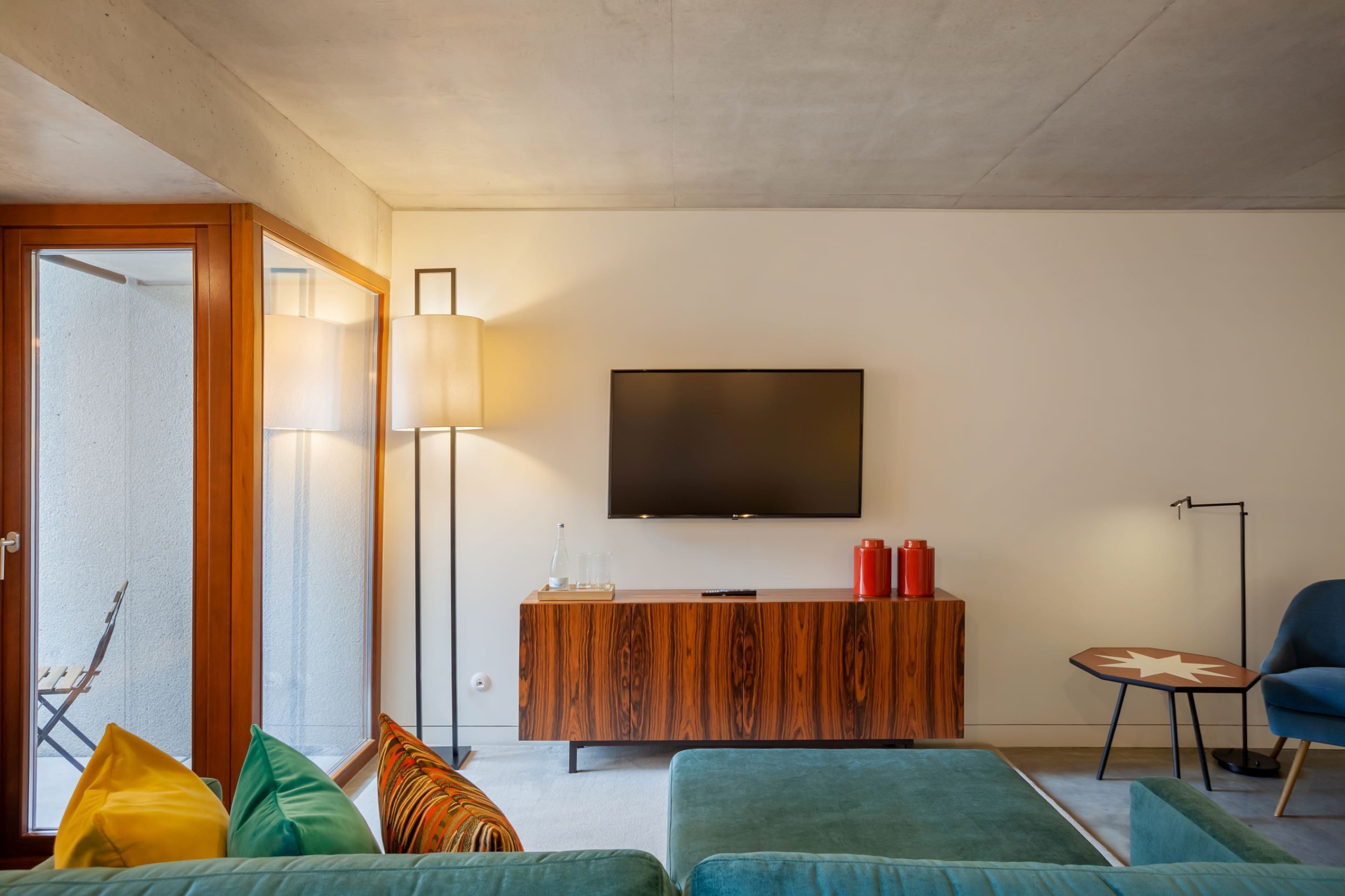 Una sala moderna con un televisor de pantalla plana grande montado en la pared, un mueble de madera con objetos decorativos, un sillón azul y un sofá con cojines de colores.