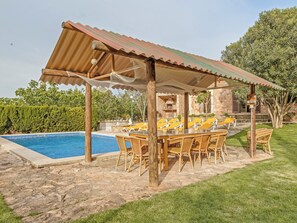 A poolside area with a covered seating area, stone flooring, and a stone building in the background.