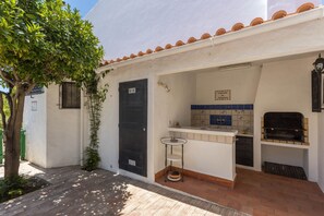 A small, white building with a tiled roof, a black door, a small outdoor kitchen area with a grill, and a tiled backsplash.