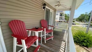 Eine Veranda mit zwei roten Adirondack-Stühlen, einem kleinen Tisch, einem Deckenventilator und Blick auf die Straße sowie die Nachbarhäuser.