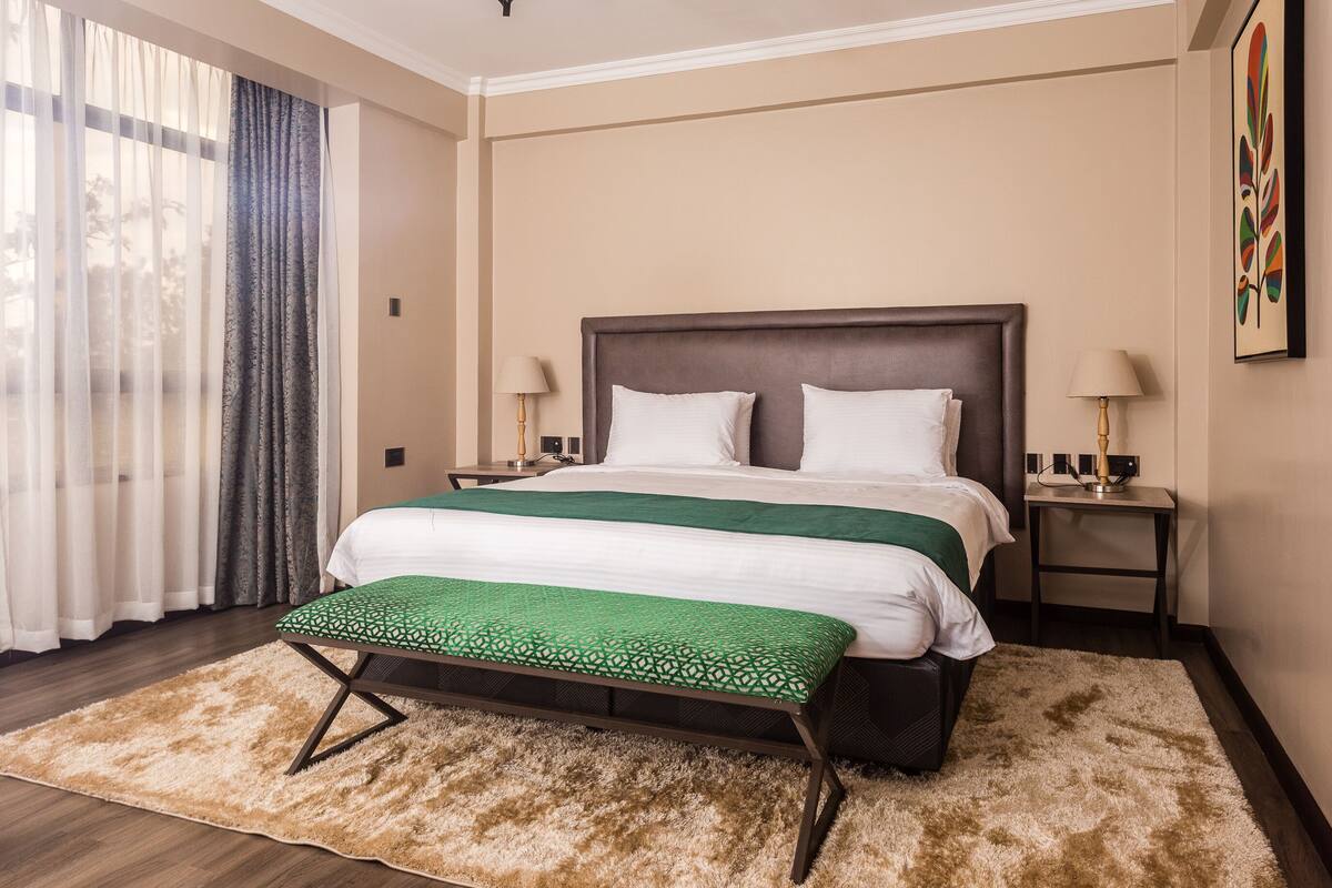 Une chambre d’hôtel avec un grand lit, deux tables de chevet, une couvre-lit verte et blanche, un banc avec un coussin à motifs verts, un tapis beige moelleux et une vue sur les arbres à travers des rideaux transparents.