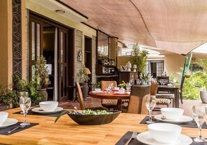Un área de comedor al aire libre con una mesa de madera preparada para dos personas, rodeada de sillas y un amplio paraguas que proporciona sombra.