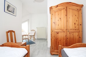 Quarto com duas camas de madeira, guarda-roupa de madeira, cadeira de vime, mesa pequena e radiador.