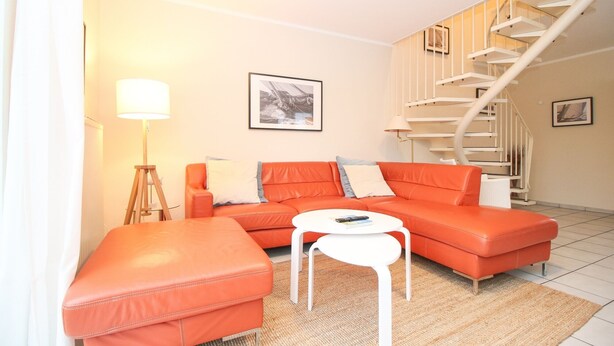 A living room with a sofa, ottoman, and a small white table. There is a staircase with a spiral design and a framed picture on the wall.