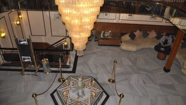 Un vaste hall d’entrée agrémenté d’un grand lustre, d’une fontaine centrale et d’un espace détente avec un canapé et une personne assise.