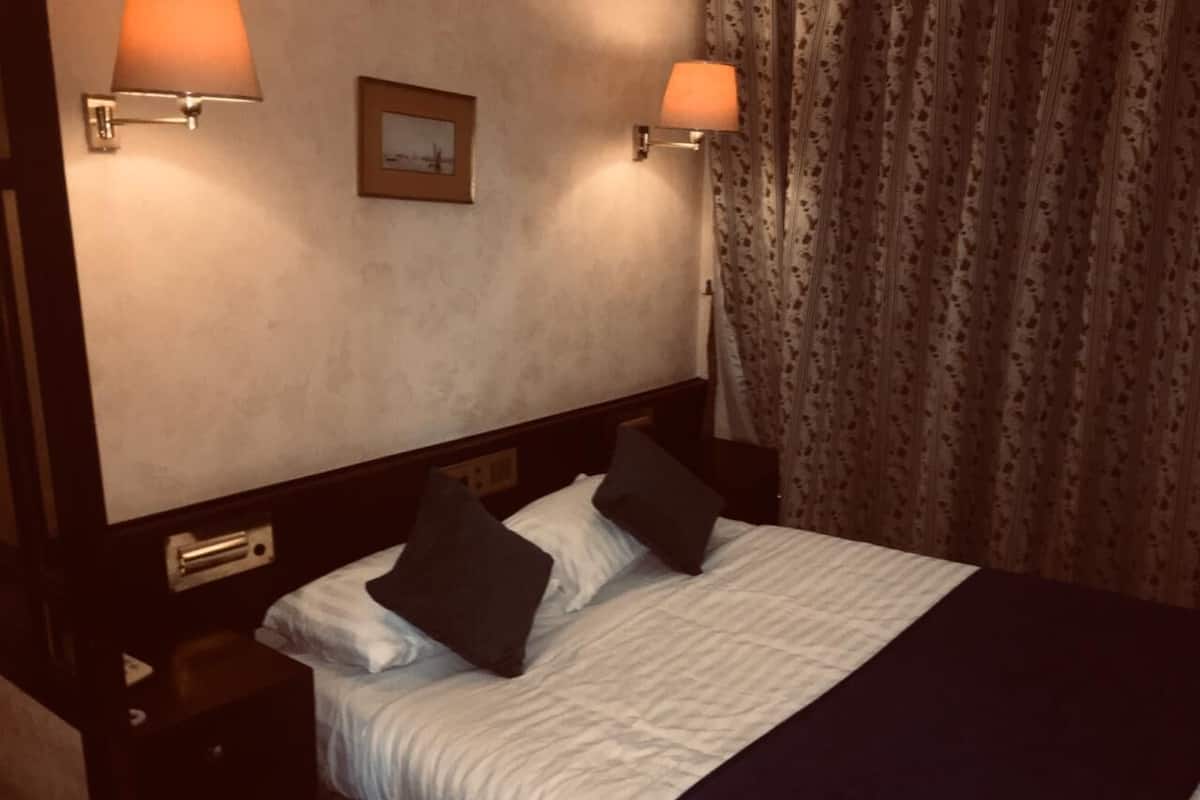A hotel room with a bed, two bedside lamps, a nightstand, a framed picture on the wall, and a curtain.