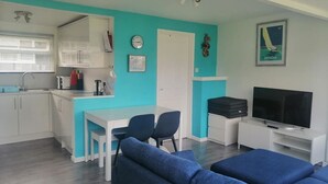 A compact living space with a kitchenette, a dining area, a seating area with blue sofas, a television, and a wall-mounted clock. - 2-bedroom sea view property 1 min from the sea & SW Coast Path (Bideford)