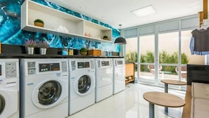 A modern laundry room with multiple washing machines, a large window with a view of greenery, and a seating area with a table.