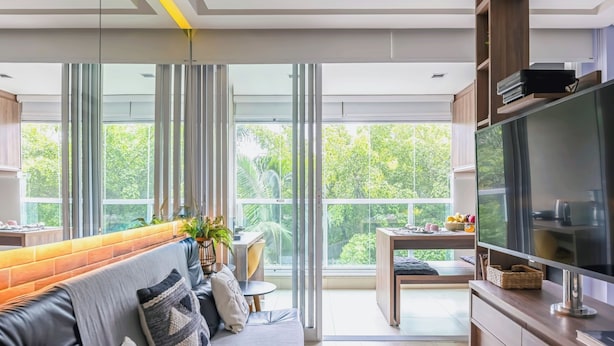A modern living room with a sofa, TV, and large windows offering a view of greenery.