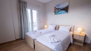 Cama doble con sábanas y toallas blancas, dos mesitas de noche con lámparas, un cuadro de un paisaje montañoso y una puerta corrediza de cristal con cortinas.