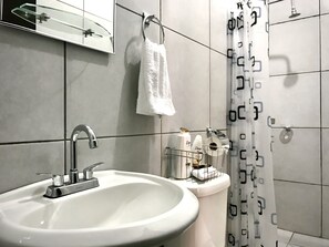 Un baño con un lavabo blanco, un espejo, una toalla colgada en un perchero y artículos de aseo en una repisa.