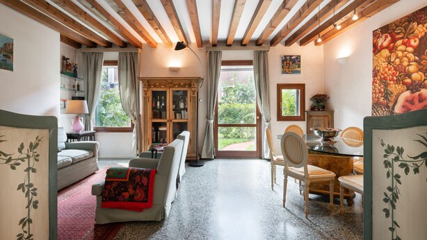 A cozy living room with a fireplace, a dining area with a table and chairs, and a view of the outdoors through large windows. - Garden Apartment (Venice)