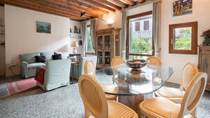 A dining area with a glass table surrounded by chairs, a wooden ceiling, and a view of greenery outside. - Garden Apartment (Venice)