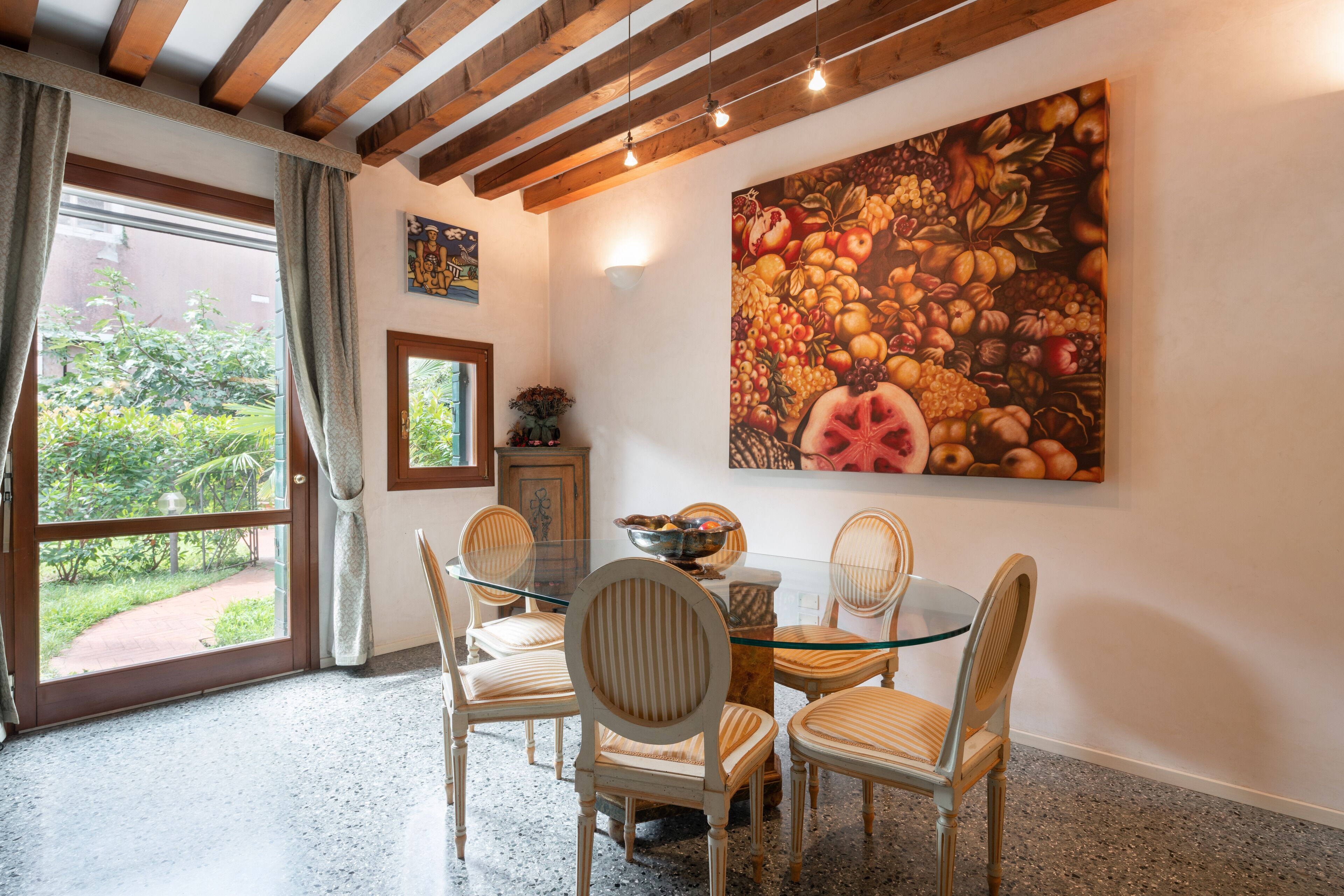 A dining area with a glass table and wooden chairs, a large fruit painting on the wall, and a view of greenery through a window.
