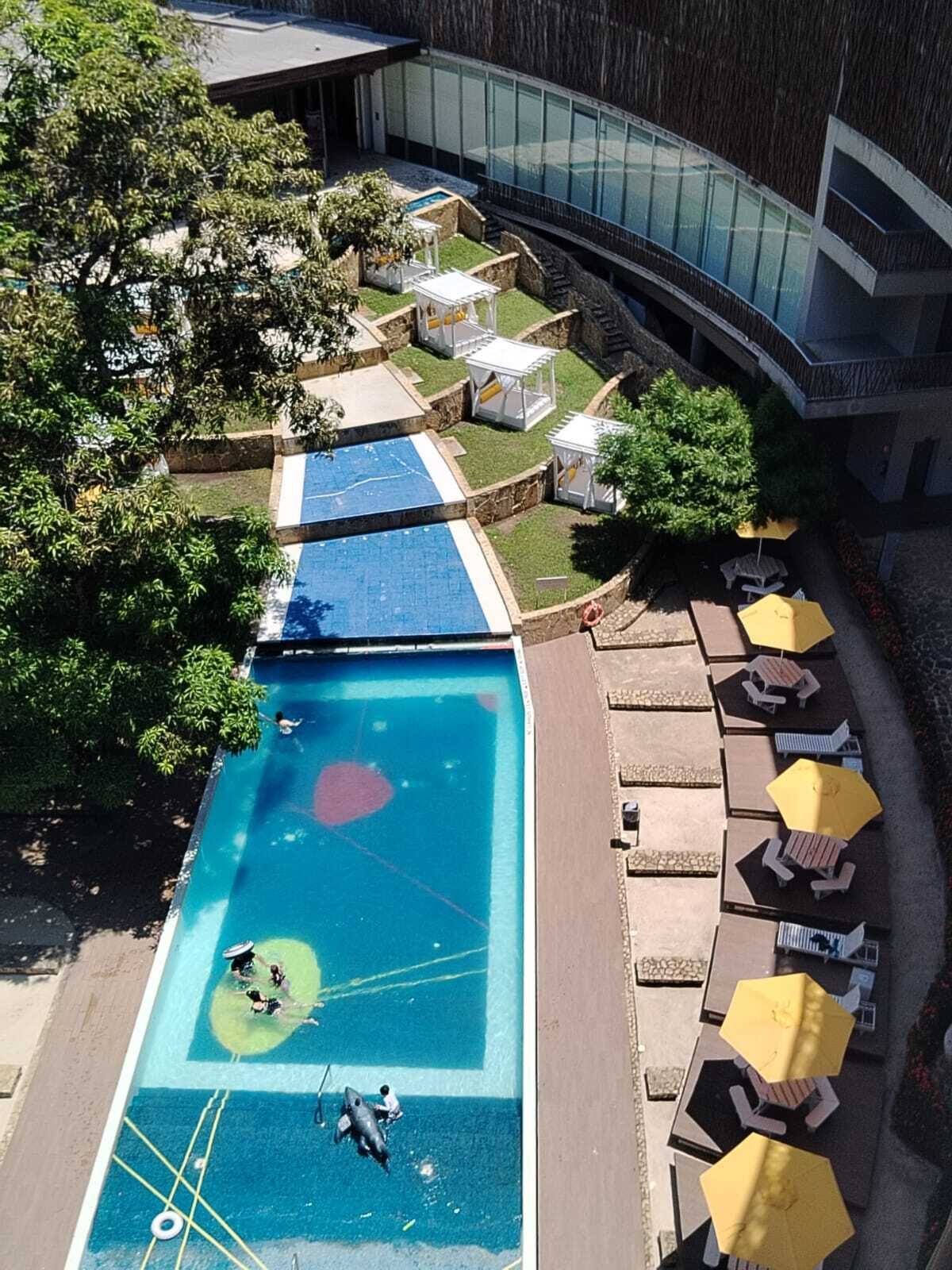 A rooftop pool with multiple levels, surrounded by lounge chairs and umbrellas, and a modern building in the background.