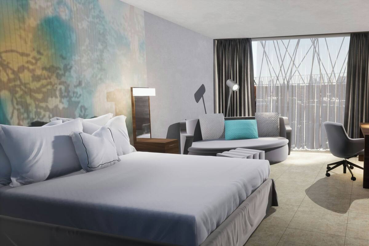 Quarto de hotel moderno com uma cama grande, uma escrivaninha, uma cadeira e vista para a cidade através de amplas janelas.