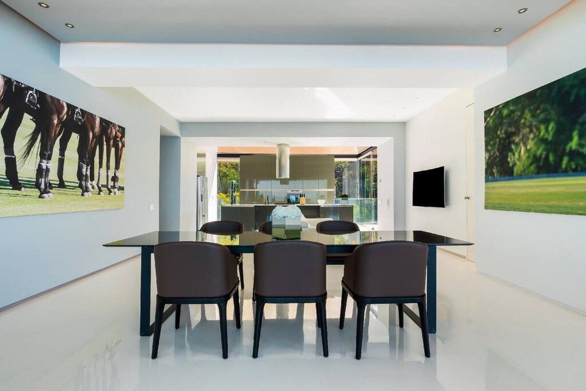 A modern dining area with a glass table and chairs, a large painting of horses on the wall, and a view into the kitchen.