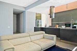 A modern living room with a beige sofa, a kitchen with dark cabinetry, and a stainless steel range hood.