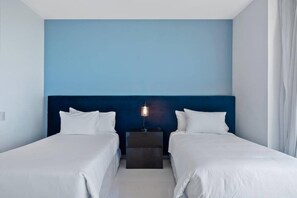 A hotel room with two beds, a dark headboard, a bedside table with a lamp, and a light blue wall.