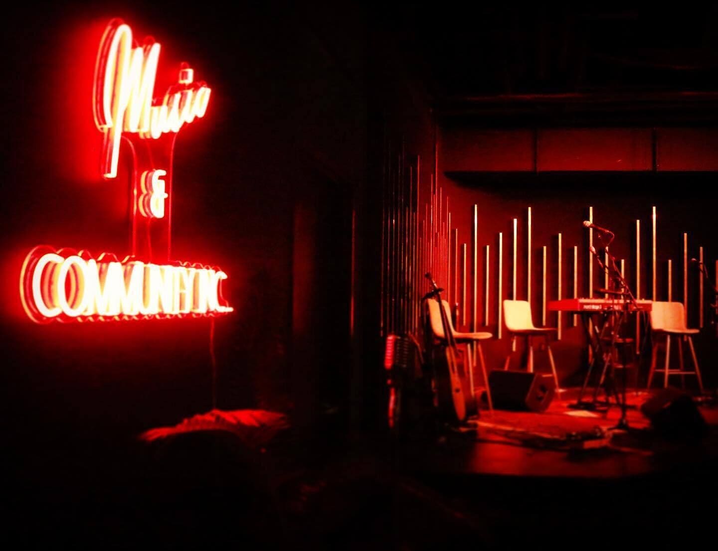 Una habitación tenuemente iluminada con un letrero de neón que dice "MUSIC COMEDY" y un escenario con instrumentos musicales y sillas.
