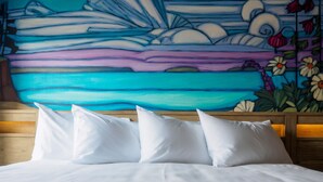 Una cama con ropa de cama y almohadas blancas, cabecera de madera y un mural colorido en la pared.