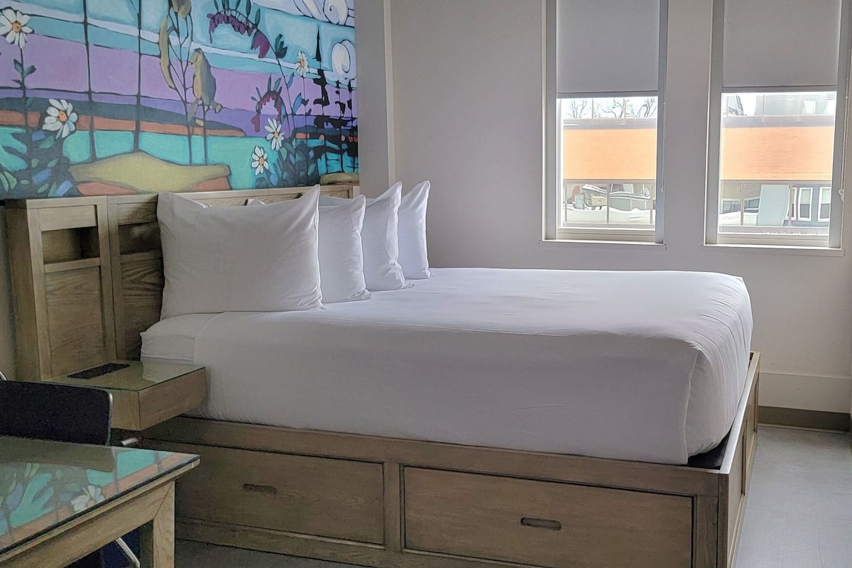 A hotel room with a bed, bedside tables, a desk, and a colorful mural on the wall.
