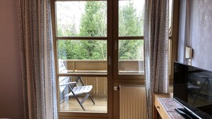 Una habitación con puerta de vidrio corredera que da a un balcón con una silla y una mesa. Hay un radiador en el suelo y un televisor sobre un mueble.